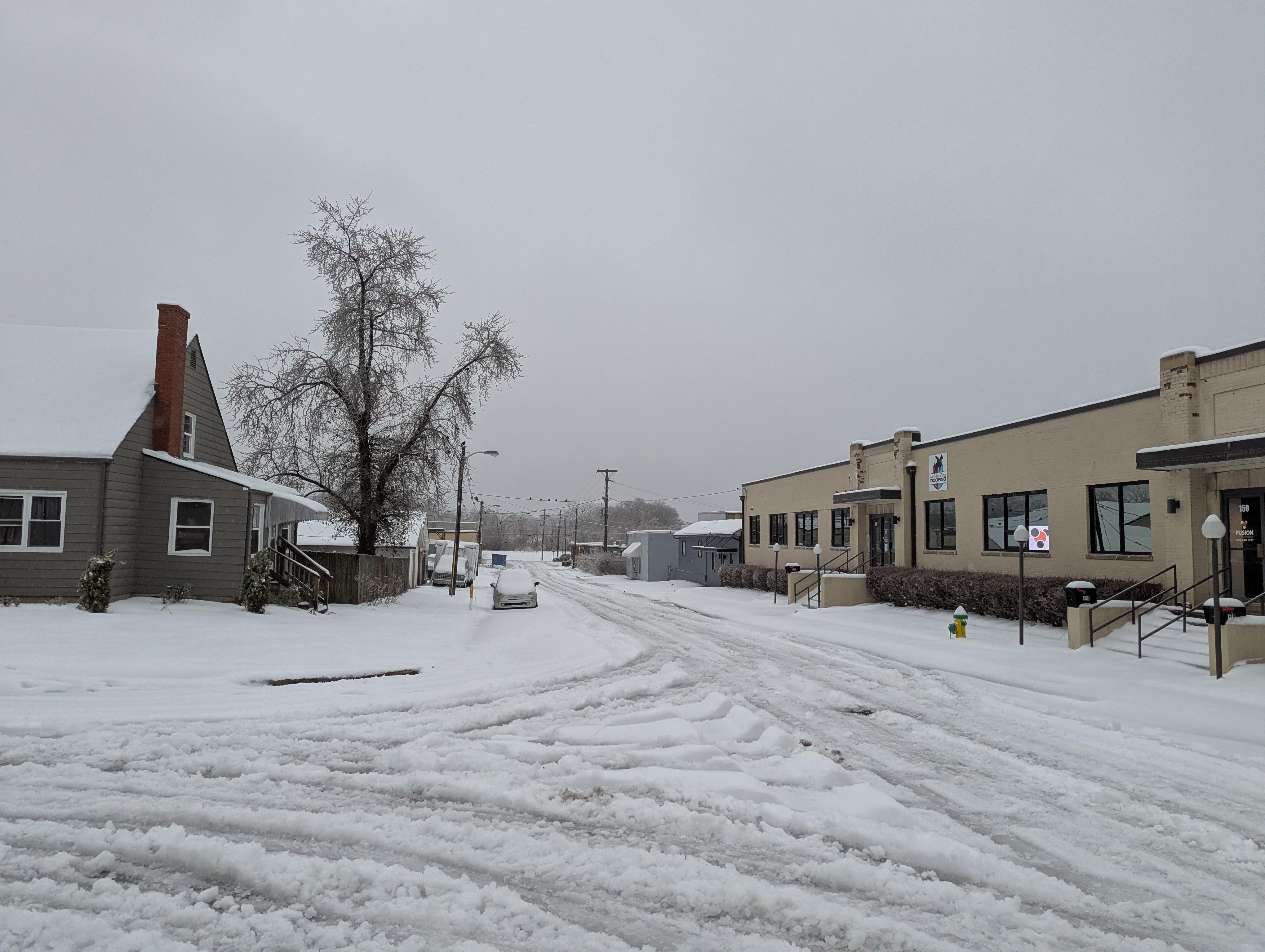 Photo of a snow-covered side street corner, with a number of tire tracks eating into the snow on the road and a near-untouched snowy sidewalk.