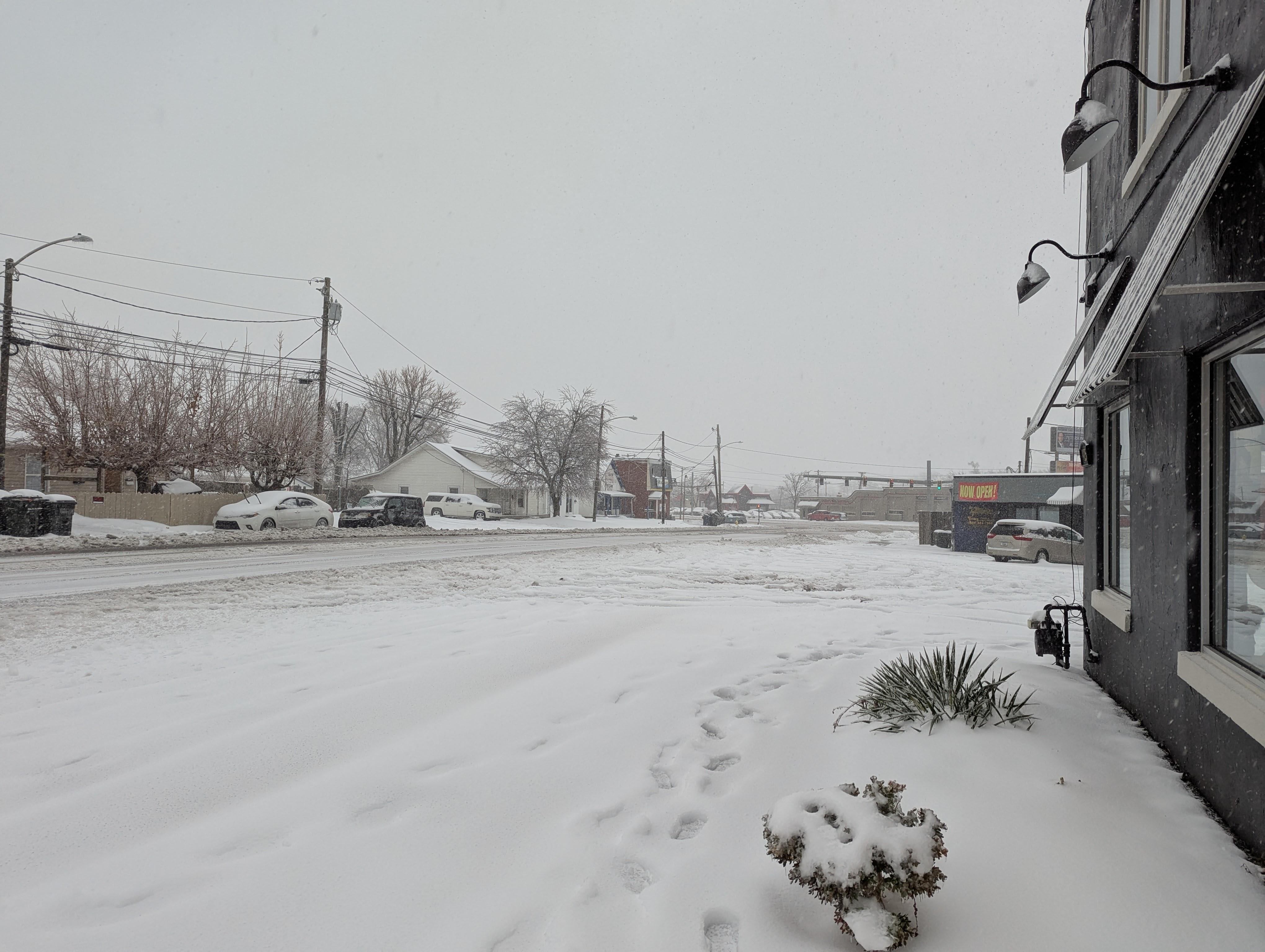 Photo of a snow-covered road, with footprints imprinting the sidewalk and a small number of snow-covered parked cars. A main road intersection and traffic light are visible in the background.