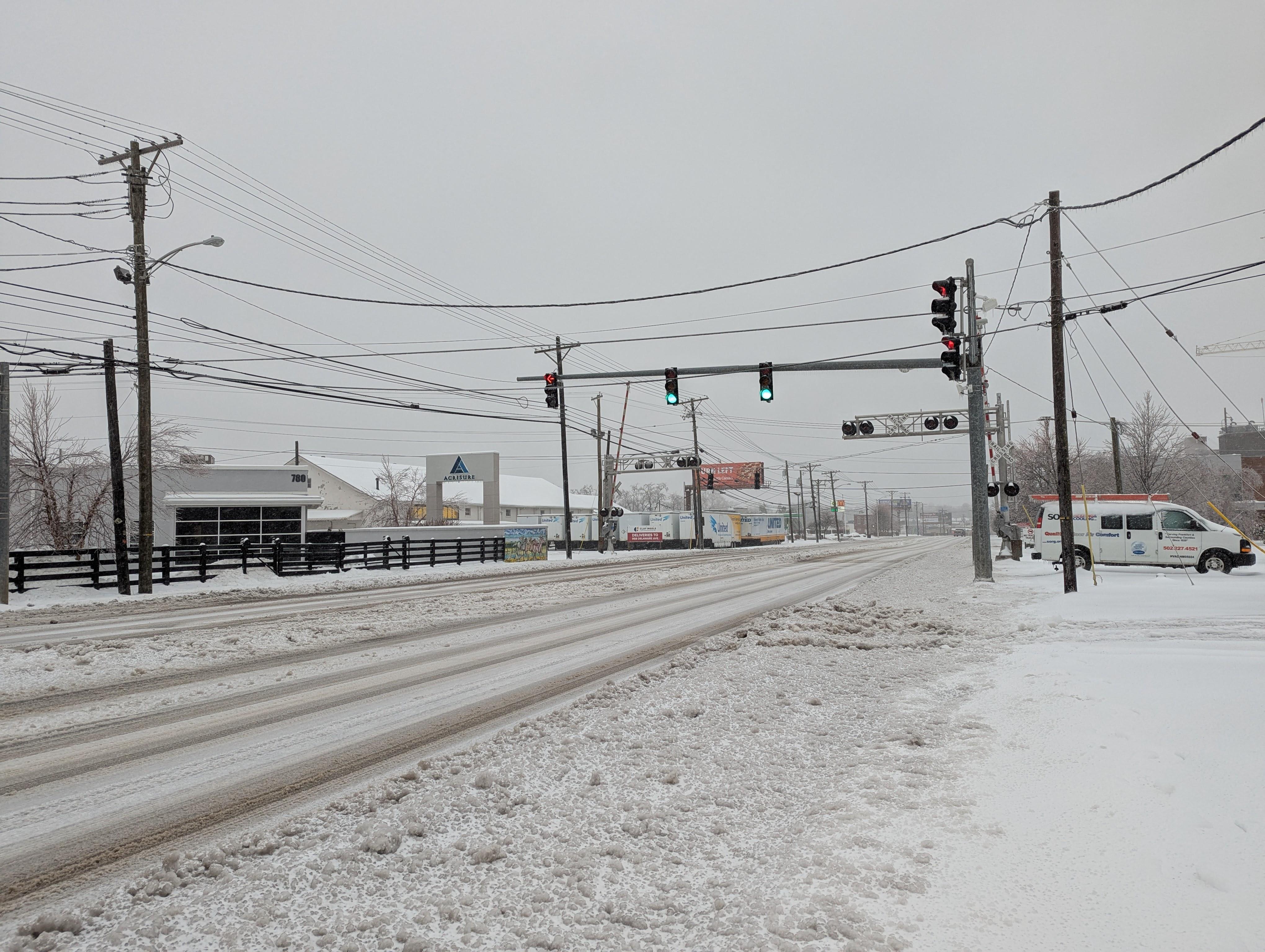 Photo of an intersection and traffic light. The road has a light amount of snow with tire tracks running through it, and the sidewalk is covered in dirty plowed snow.