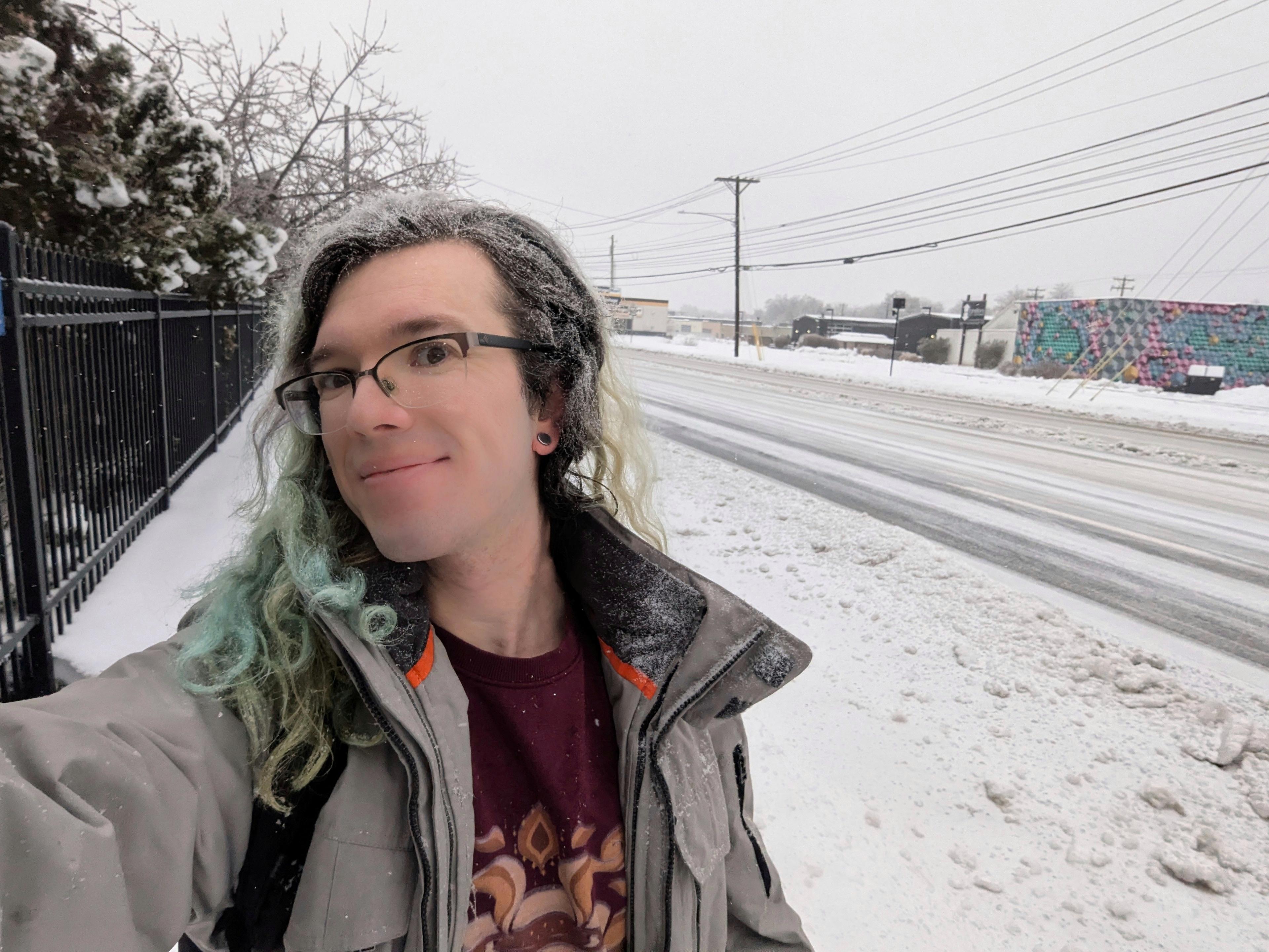 Selfie of me smiling and walking alongside a snowy sidewalk, with a lightly-covered street and power lines in the background. My hair is dusted with snow and I am wearing a tan winter jacket.