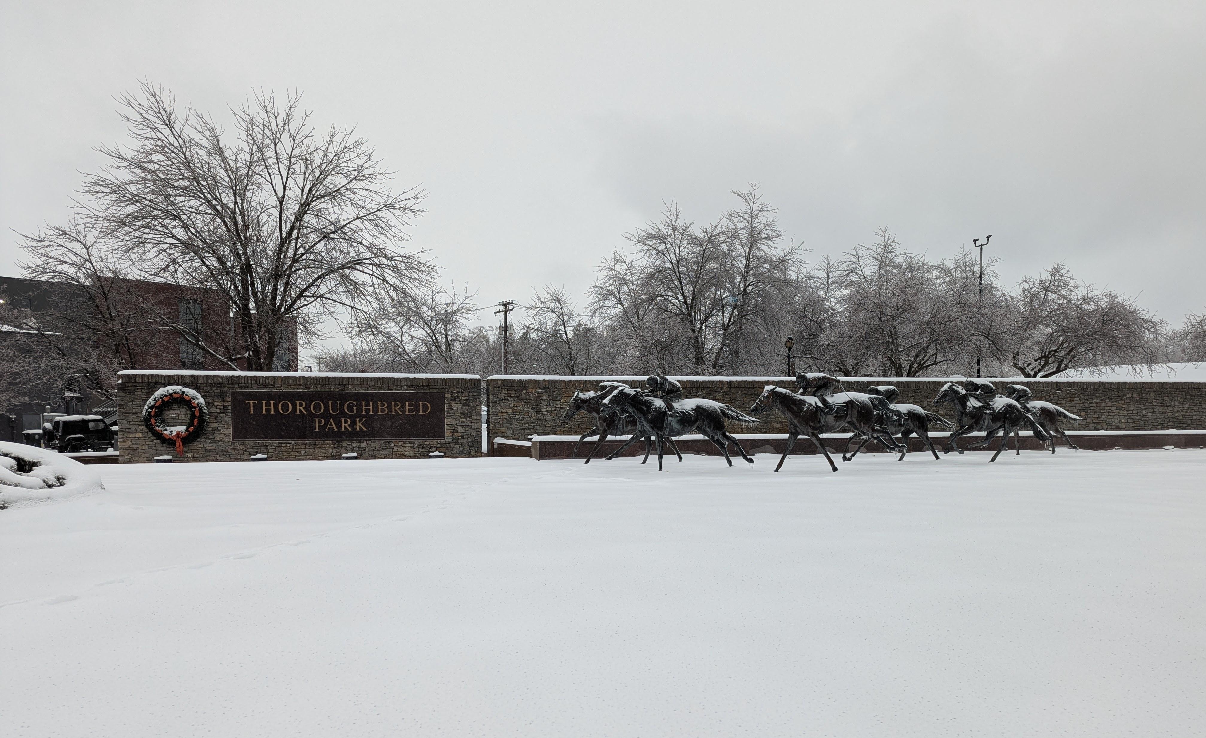 Photo of a park entrance, with a sign reading "Thoroughbred Park" on a stone wall. A group of statues of running horses are to the right of the sign and coated in a layer of snow. The foreground is almost entirely untouched snow, and the background is lined with bare and icy trees.